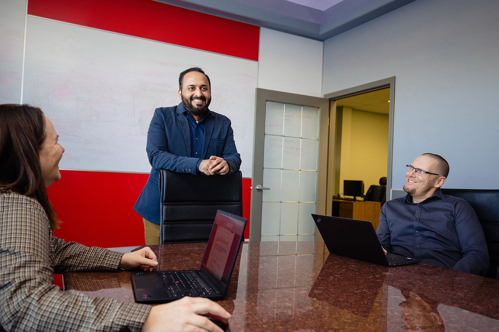 A man stands at the head of a boardroom table as two of his colleagues look up at him.